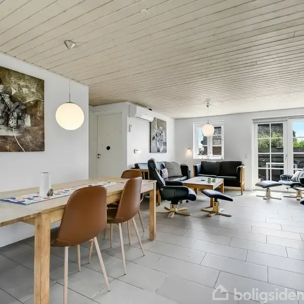 Et lyst stueområde med spisebord og stole i forgrunden, lænestole og sofa mod vinduerne i baggrunden, og dekoreret med kunst på væggene.
