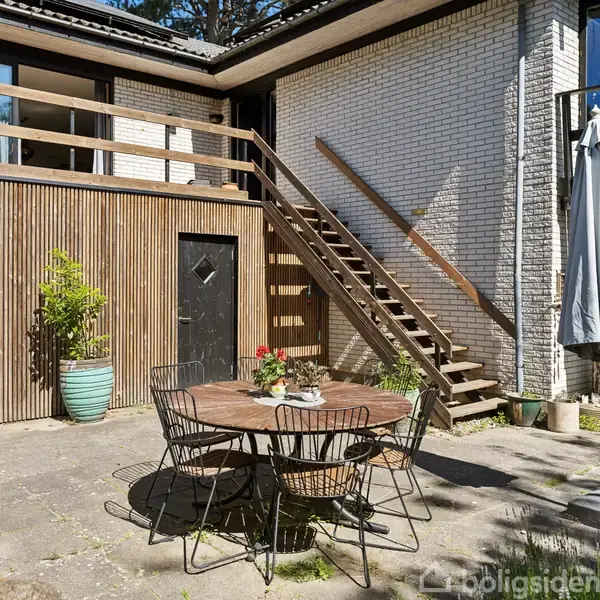 Rundt havebord med stole står på en stenbelagt terrasse i solen. Trætrappe fører op til altan på hvid murstensbygning. Store blomsterkrukker og parasol omgiver området.