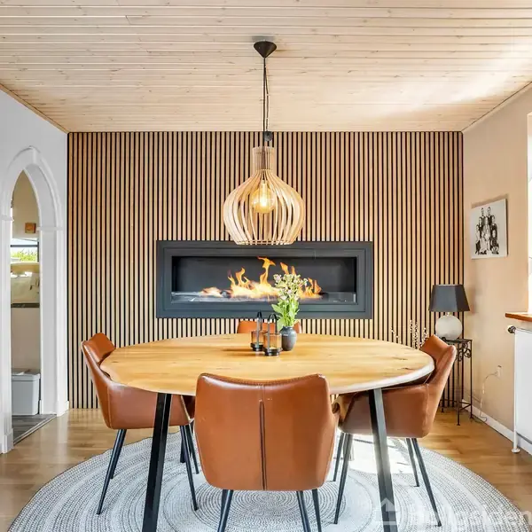 Rundt bord med brune stole står foran en moderne, vægmonteret pejs i et rum med træpanelvæg og træloft. En stor lampe hænger over bordet, med dekorationer omkring.