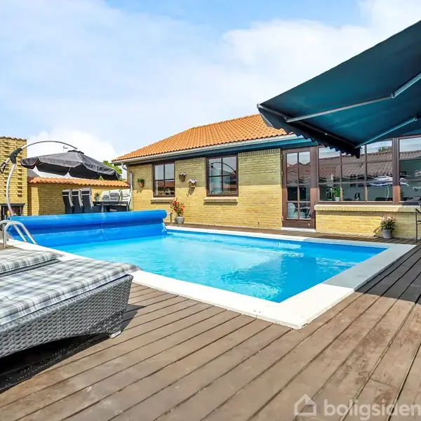 En swimmingpool med blåt vand ligger stille på en træterrasse, omgivet af liggestole og parasoller. En gul murstensbygning med rødt tag danner baggrunden.