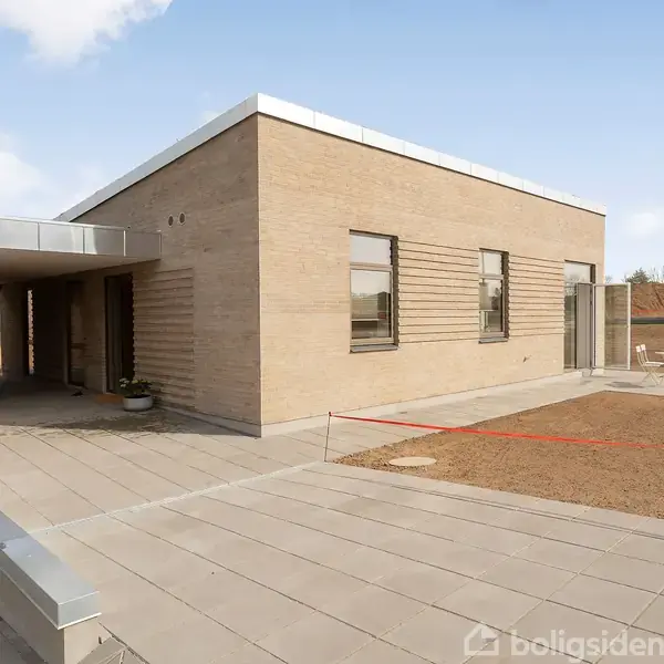 En moderne bygning med fladt tag står på en stor, flisebelagt terrasse. Bygningen har lyse mursten og store vinduer. Omgivelserne er åbne med en græsplæne under etablering.