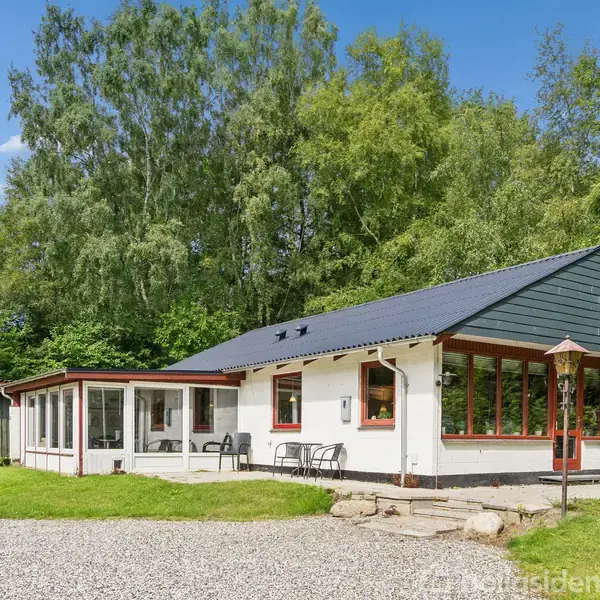 Et hvidt hus med sort tag står omgivet af grønne træer. To stole er på en lille terrasse ved huset, og en grusvej fører op til indgangen.