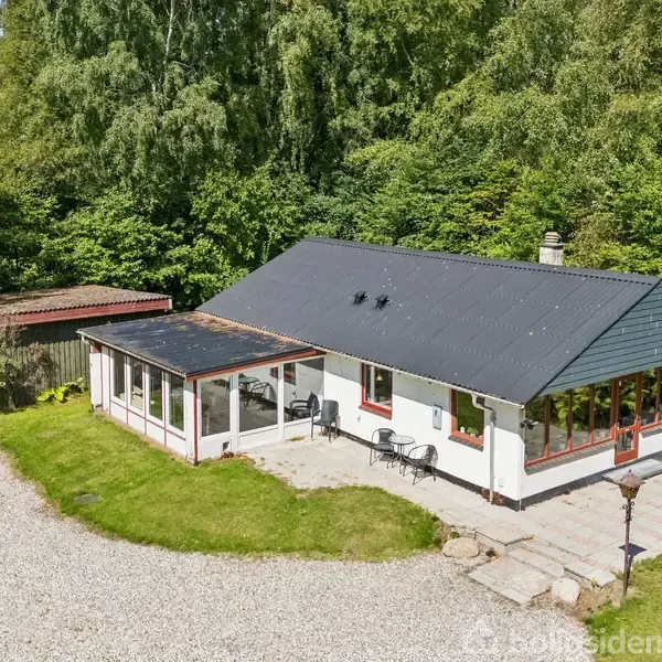 Hus med sort tag og hvide vægge, står stille omgivet af grøn vegetation. Til højre er der havemøbler, og til venstre en lille udestue. Grusbelagt indkørsel foran.