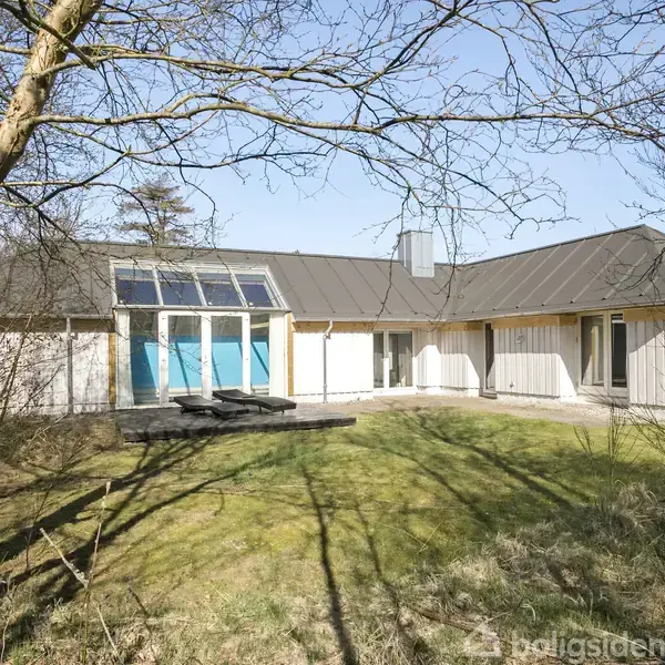 Et moderne, hvidt hus med store vinduer og gråt tag ligger i en åben have med græsplæne. Solen skinner, og omgivelserne har spredte træer uden blade.