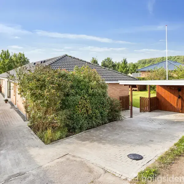 En villa med carport er omgivet af hække og buske. Fliser fører op til huset i et grønt, landligt område. Himlen er klar med få skyer.