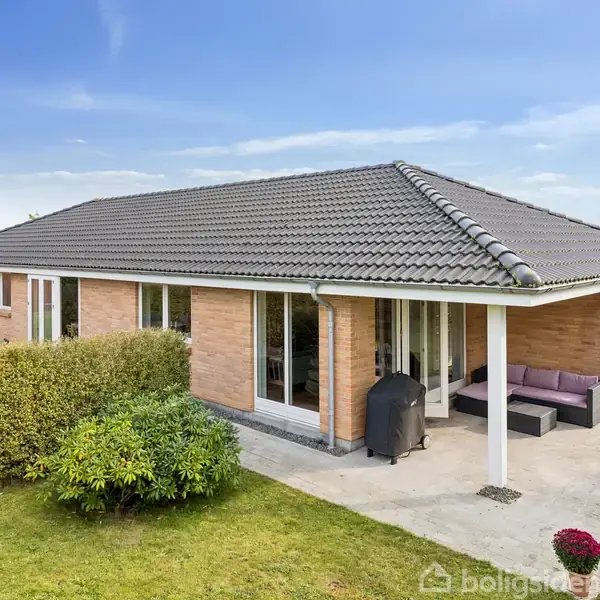 Et hus med tegltag står stille i en have med en flisebelagt terrasse. Omkring terrassen er der tætte buske og en græsplæne. En sofa og grill ses under overdækningen.