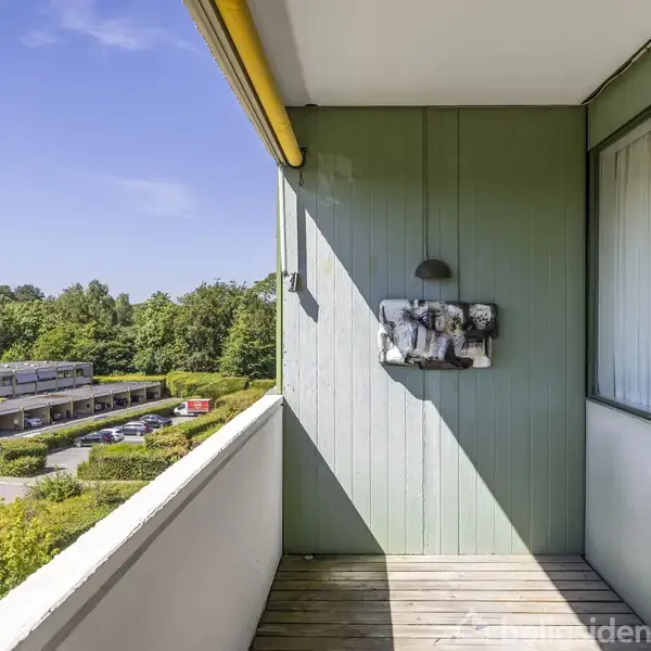 En altan med træplanker, hvor en lys lampe hænger på væggen. I baggrunden ses et boligområde med parkeringspladser, træer og blå himmel.
