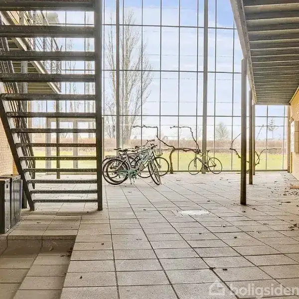 En metaltrappe står stille i en overdækket, åben gård med flisegulv og gule murstensvægge. Flere cykler er parkeret ved glasvægge, der giver udsigt til et grønt landskab.
