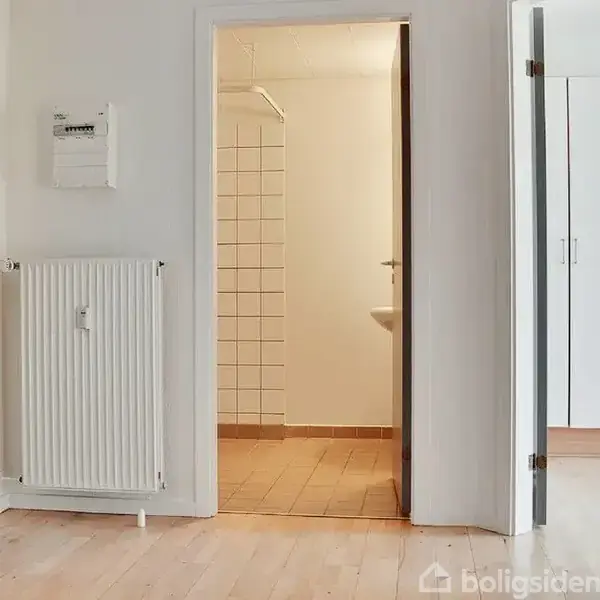 Radiator og dør: Radiatoren hænger på væggen ved siden af en åben dør, der fører til et badeværelse med fliser. Trægulve løber gennem rummet, der har lyse vægge.