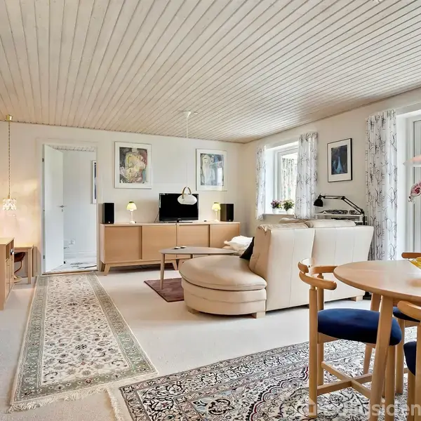 En stue indeholder en rund sofa og et fjernsyn på et skab. Der er tæpper på gulvet, spisebord med tre stole og blomster. Rummet har store vinduer og lys træbeklædning.