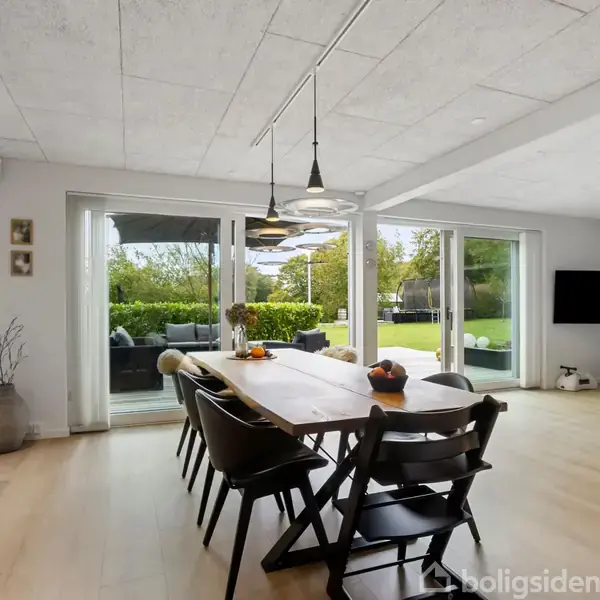 Spisebord med seks stole står i et lyst rum. Rummet har store vinduer med udsigt til en grøn have. Lamper hænger over bordet, og der er en vægmonteret tv-skærm.