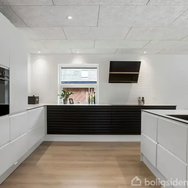 Et moderne køkken med hvide skabe og en sort emhætte. Rummet har trægulv, et vindue i midten, og en dekorativ plakat af et farverigt hjortehoved på væggen.