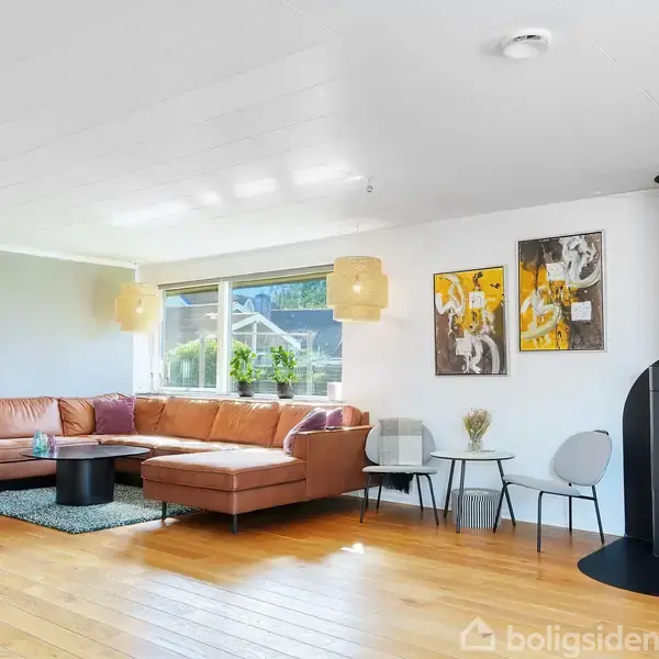 En stue med en stor brun lædersofa og et brændende sort brændeovn. I vindueskarmen står planter. Kunstværker hænger på de hvide vægge.