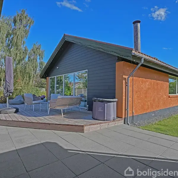 Et moderne hus står med træterrasse, hvor havemøbler er arrangeret til afslapning. Terrasseområdet er omkranset af en velplejet græsplæne, og himlen er klar og blå.
