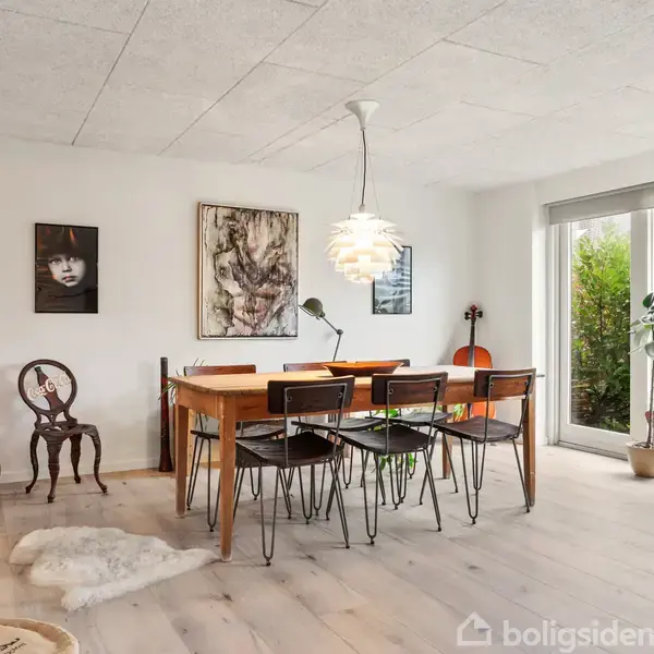 Et spisebord med seks stole står midt i et lyst rum. Der er kunstværker på væggen og en lampe hængende over bordet. Et stort vindue viser udsigt til grønne buske.
