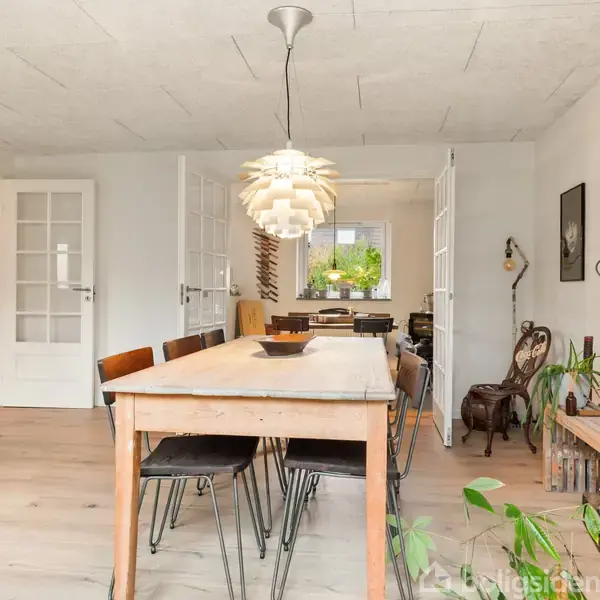 Spisebord med mange stole står midt i et lyst rum. En moderne lampe hænger ned over bordet. Væggene er dekoreret med kunst og planter, hvilket skaber en hyggelig atmosfære.