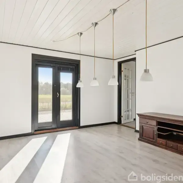 Stue med hvide vægge og loft, trægulv, fire loftslamper, og en mørk træreol. Store vinduer og glasdør giver udsyn til en græsplæne.