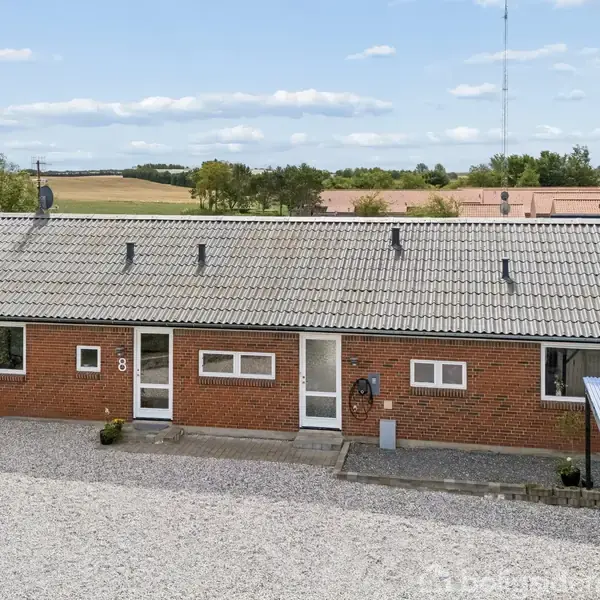 En rød murstensbygning med gråt tag står foran en grusbelagt gårdsplads. Tallet "8" er synligt ved siden af døren. Landskab og bygninger er i baggrunden.