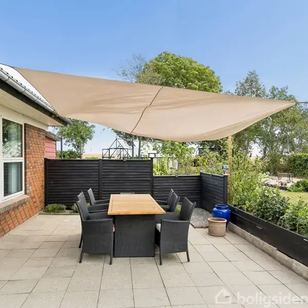 Terrassebord med seks sorte stole står under en stor beige solsejl. Terrassen har flisebelægning, omgivet af planter, redskabsskur og en græsplæne med træer i baggrunden.