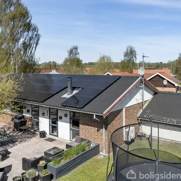 Et moderne hus med solpaneler på taget står i en have med terrasse, havemøbler og en trampolin. Træer omgiver ejendommen, og blå himmel ses i baggrunden.