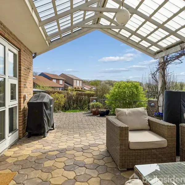 Overdækket terrasse med kurvemøbler i et boligområde. Klart glasloft tillader udsigt til blå himmel, huse og grønne omgivelser. En grill står ved siden af en murstensvæg.