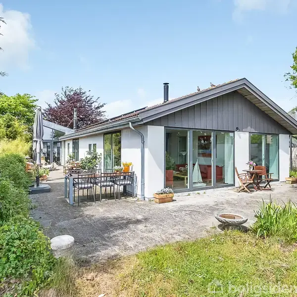 Huset står i en grøn have med store vinduer og terrasse, hvor to borde og stole er placeret. Havemøblerne er omgivet af planter og træer i et solrigt område.