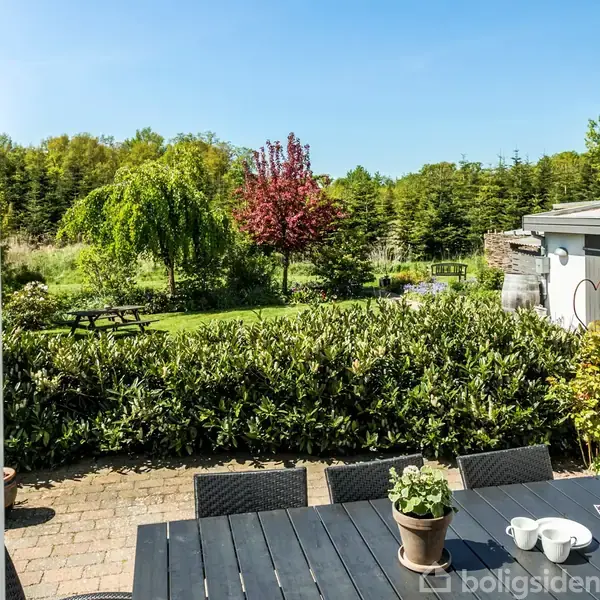 Et sort havebord med kaffekopper står på en terrasse omgivet af grønne buske. I baggrunden ses en have med træer og en bænk under en lys blå himmel.