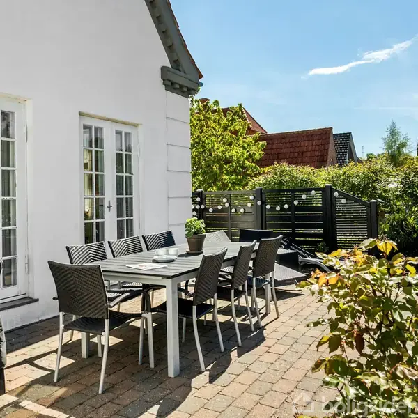 Terrassebord med stole står på flisebelagt område i solskinsvejr ved siden af et hvidt hus. Omkring er der grønne planter og en grill, med et hegn bagved.