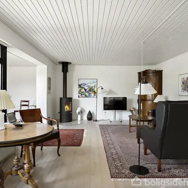 Stue med pejseovn til venstre i hjørnet. Rundt bord med lampe står foran stort vindue. Sofa og lænestol ved siden af tv. Tæppe på gulvet.