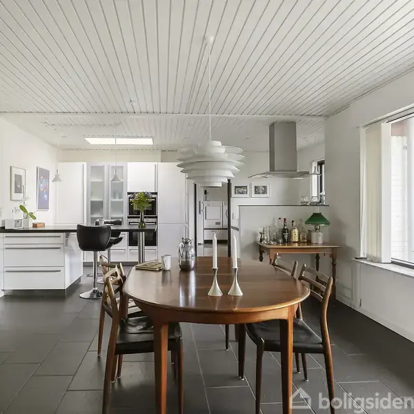 Rundt spisebord med stole, dækket med lys, står i en åben moderne stue-køkken. Baggrunden viser et hvidt køkken med barstol, mens et stort vindue giver lys til rummet.