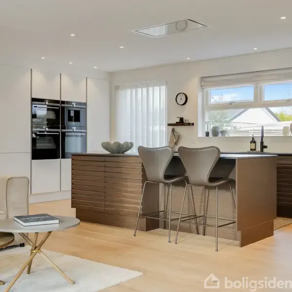 Moderne køkken med en stor ø, to barstole og integrerede ovne til venstre. Et sofabord med blomster på en lys trægulv i en stilfuld stue.