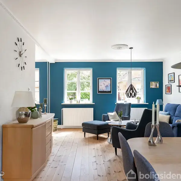 Stue med blå sofa og lænestol, belyst af naturligt lys fra vinduer. Spisebord med stole forrest og vægge dekoreret med billeder. Indbydende atmosfære med trægulve.