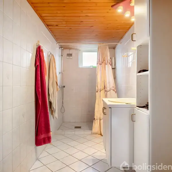 Badeværelse med hvidt flisegulv og vægge, træloft, brusebad bagerst, håndvaske og skabe til højre, farverige håndklæder hænger til venstre ved toiletpapirholder.