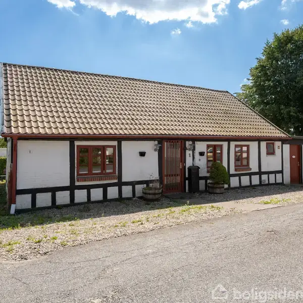 Et hvidt bindingsværkshus med brunlige detaljer står ved en stille vej. Huset har et tegltag og vinduer med røde rammer. Det er omgivet af grønne træer og en græsplæne.