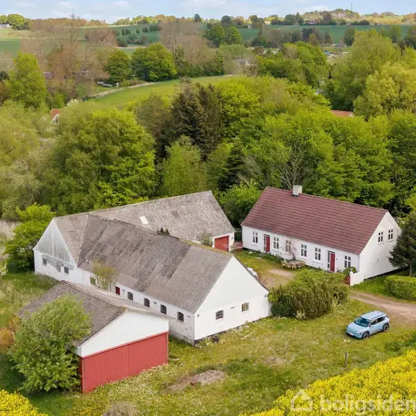 En traditionel dansk gård med hvid facade og rødt tag, omgivet af frodig grøn vegetation og marker. En blå bil holder parkeret foran bygningerne under en klar himmel.