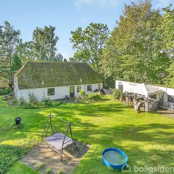 Huset har et stråtækt tag og ligger blandt frodige træer og buske. Foran huset er en græsplæne med en gynge og et blåt plastikbassin.