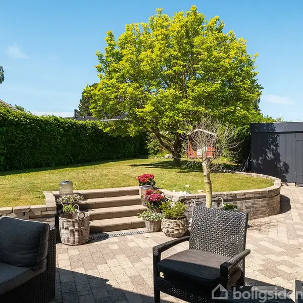 Terrassemøbler står på en flisebelagt terrasse. En trappe fører op til en plæne med en stor, grøn træ midt i en privat have omkranset af hække og bygninger.