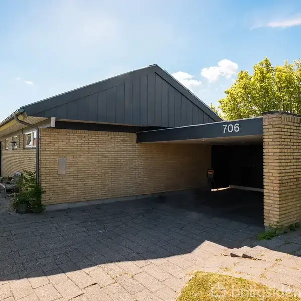 En énstok villa af mursten med et skråt tag har en overdækket carport, hvor nummeret "706" er synligt. Carporten står ved siden af et hus med en flisebelagt indkørsel.