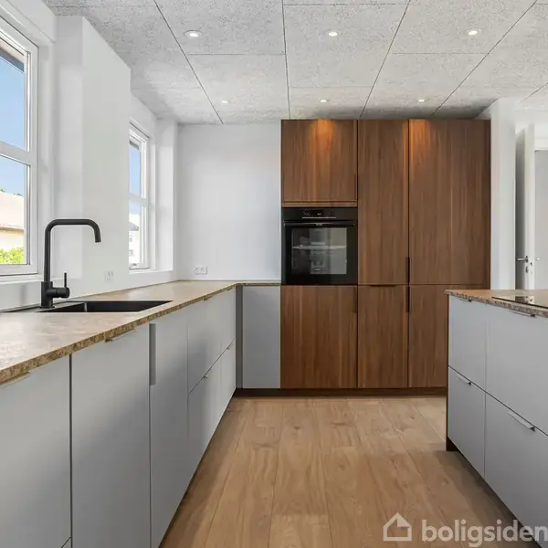 Køkken med moderne design, hvor en ovn er integreret i en høj, trælignende skabssektion. Lysegrå skabe og mørke bordplader i et rum med stort, naturligt lysindfald fra vinduerne.