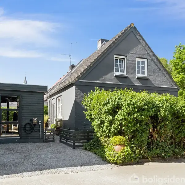 Et gråt hus med skråt tag står ved siden af en garage omgivet af grønne buske. Sollyst område med blå himmel. Der er ingen tekst i billedet.