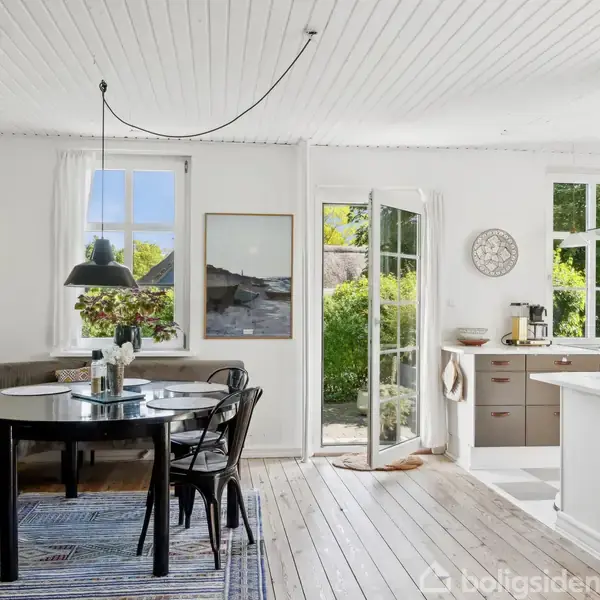 Et åbent køkken-alrum med et rundt, sort spisebord og stole på et stribet tæppe. Til venstre er en dør åben til en grøn have. Hvide vægge og lofter.