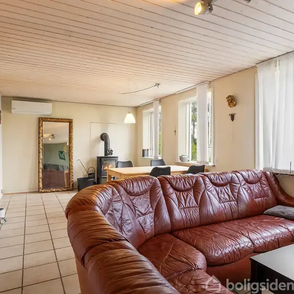 En stor brun lædersofa står i en stue med lyse fliser på gulvet. Langs væggen er der tre vinduer med persienner. Et spisebord står i baggrunden ved siden af et spejl og en brændeovn.