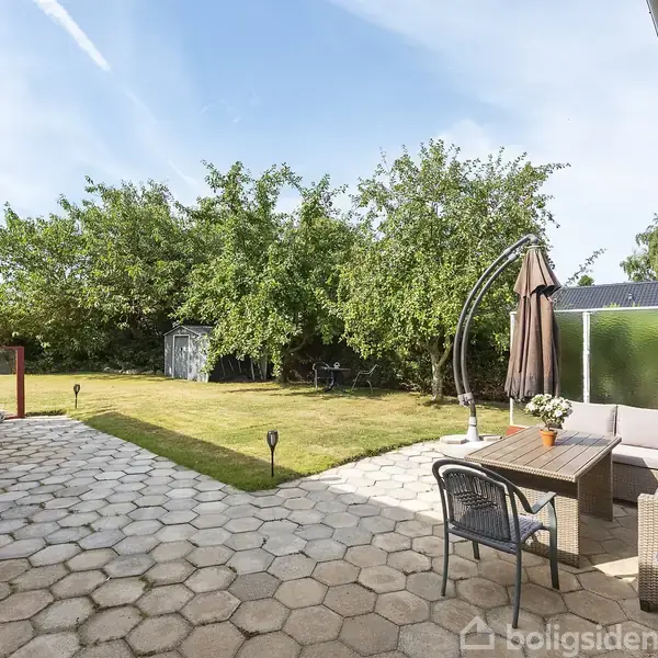 Terrasse med møbleret hjørnesofa og bord under en stor parasol. Handlingen viser en fredfyldt have med grønne træer i baggrunden, omgivet af en velplejet græsplæne og flisebelagt gulv.