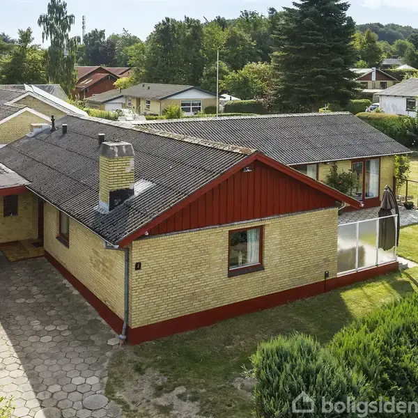 Et gult murstenshus med rødt træværk og garage, omgivet af en have med buske. Taget er skråt, og der er en skorsten. Beliggende i et villakvarter med flere huse.