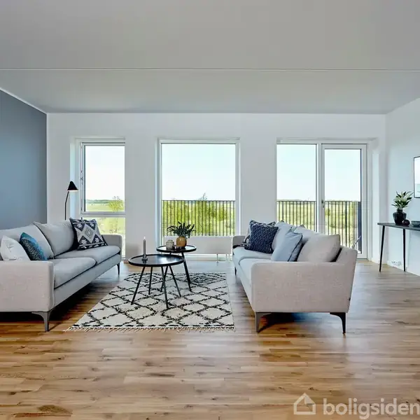To sofaer står overfor hinanden med et sofabord imellem i en moderne stue. Store vinduer giver naturligt lys, og gulvet er af lyst træ.