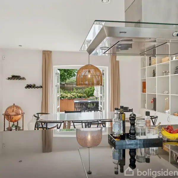 En moderne køkkenø i et stilfuldt køkken er dækket med krydderier og frugt i skåle. I baggrunden er der en spisestue med stole og en åben dør mod en terrasse.