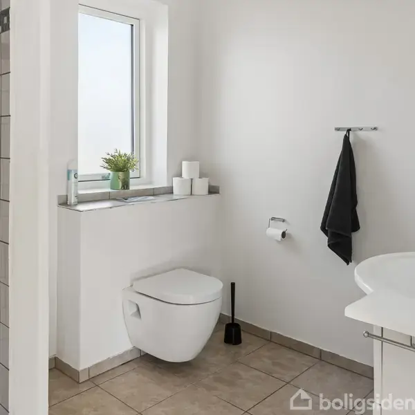 Toilet hænger på væggen; til venstre er en flisebelagt bruseniche. På vindueskarmen står en plante og toiletpapir. Håndklæde hænger på væggen i et lyst og enkelt badeværelse.