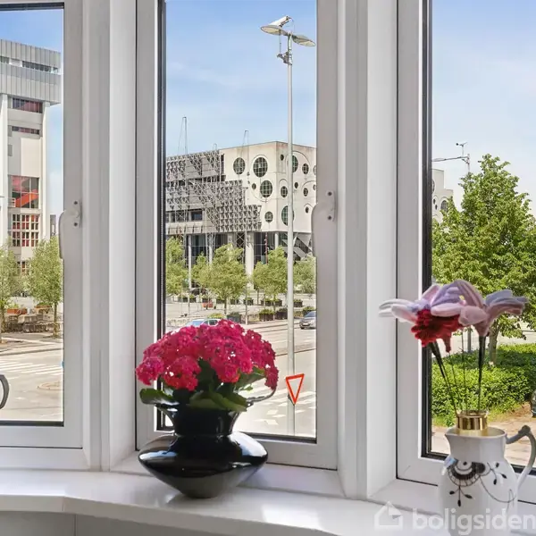 Et vindue med tre sektioner, indeholder en sort vase med lyserøde blomster og to kander. Udsigten viser en byvej med træer, biler og bygninger, inklusive "NORDKRAFT."