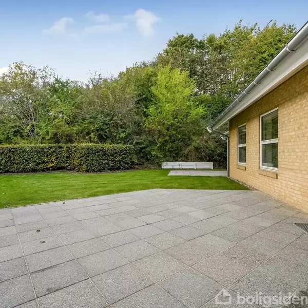 En flisebelagt terrasse ligger ved et gult murstenshus med store vinduer; i baggrunden er der en grøn græsplæne og frodig hæk omringet af træer under en blå himmel.