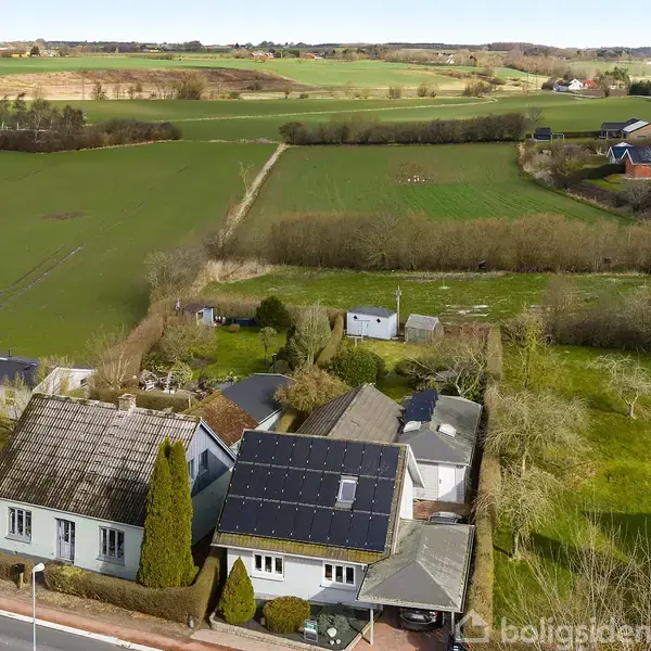 Huse med solpaneler ligger langs en vej, omgivet af træer og haver. Landskabet i baggrunden består af åbne marker og spredte huse under en klar himmel.